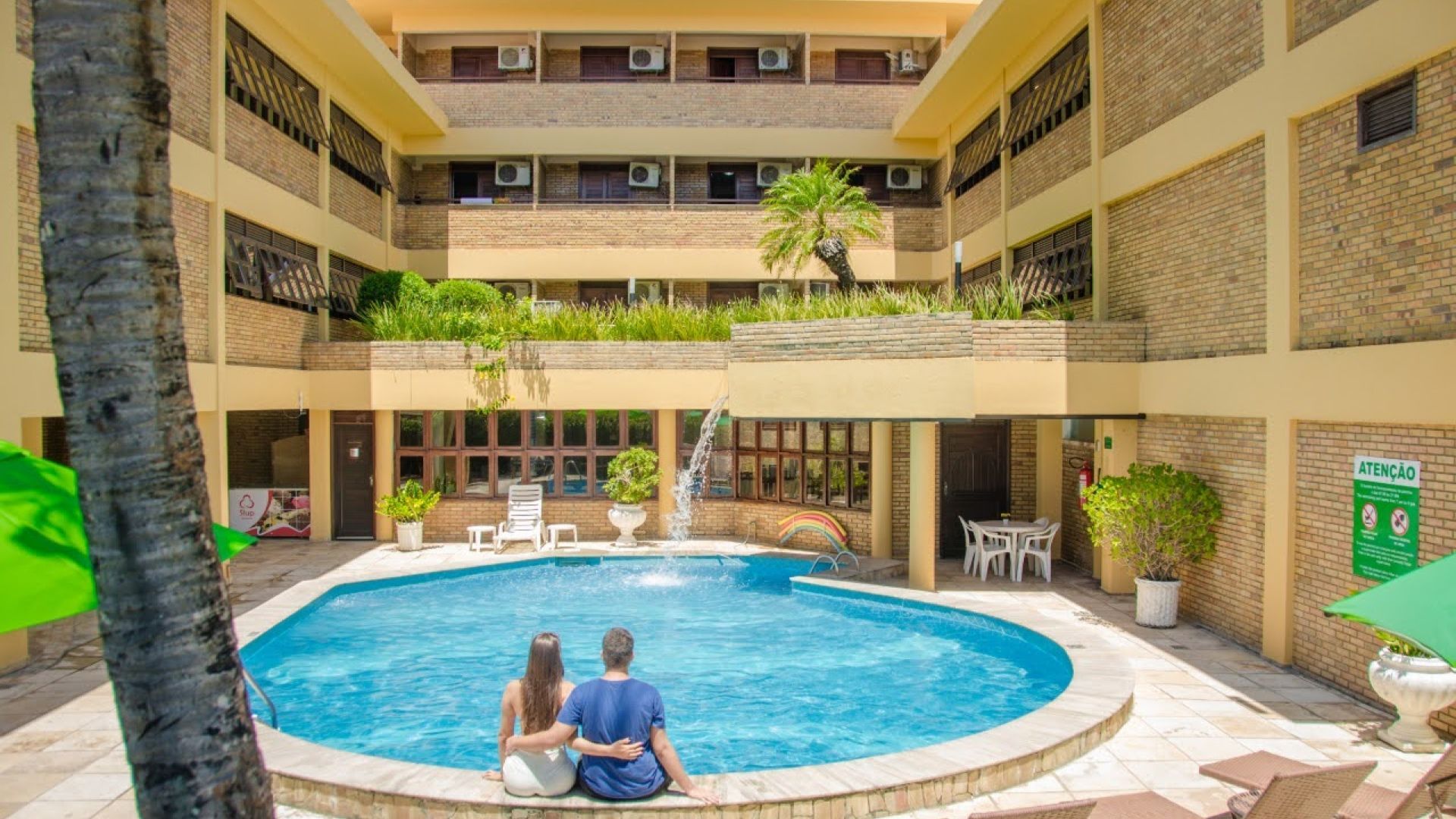 Um homem e uma mulher estão sentados na borda de uma piscina. - Pizzato Praia Hotel