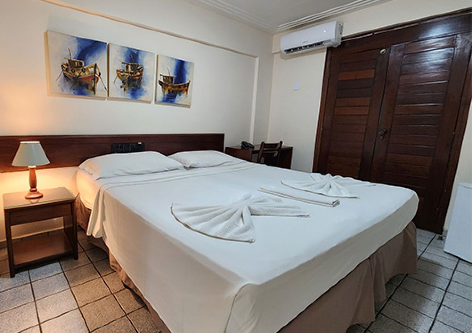 Um quarto de hotel com uma cama, mesa de cabeceira, abajur e pinturas na parede. - Pizzato Praia Hotel