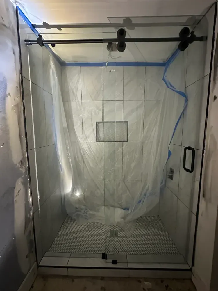 Empty shower stall enclosed in clear plastic, with a tiled floor and blue painter’s tape along the edges