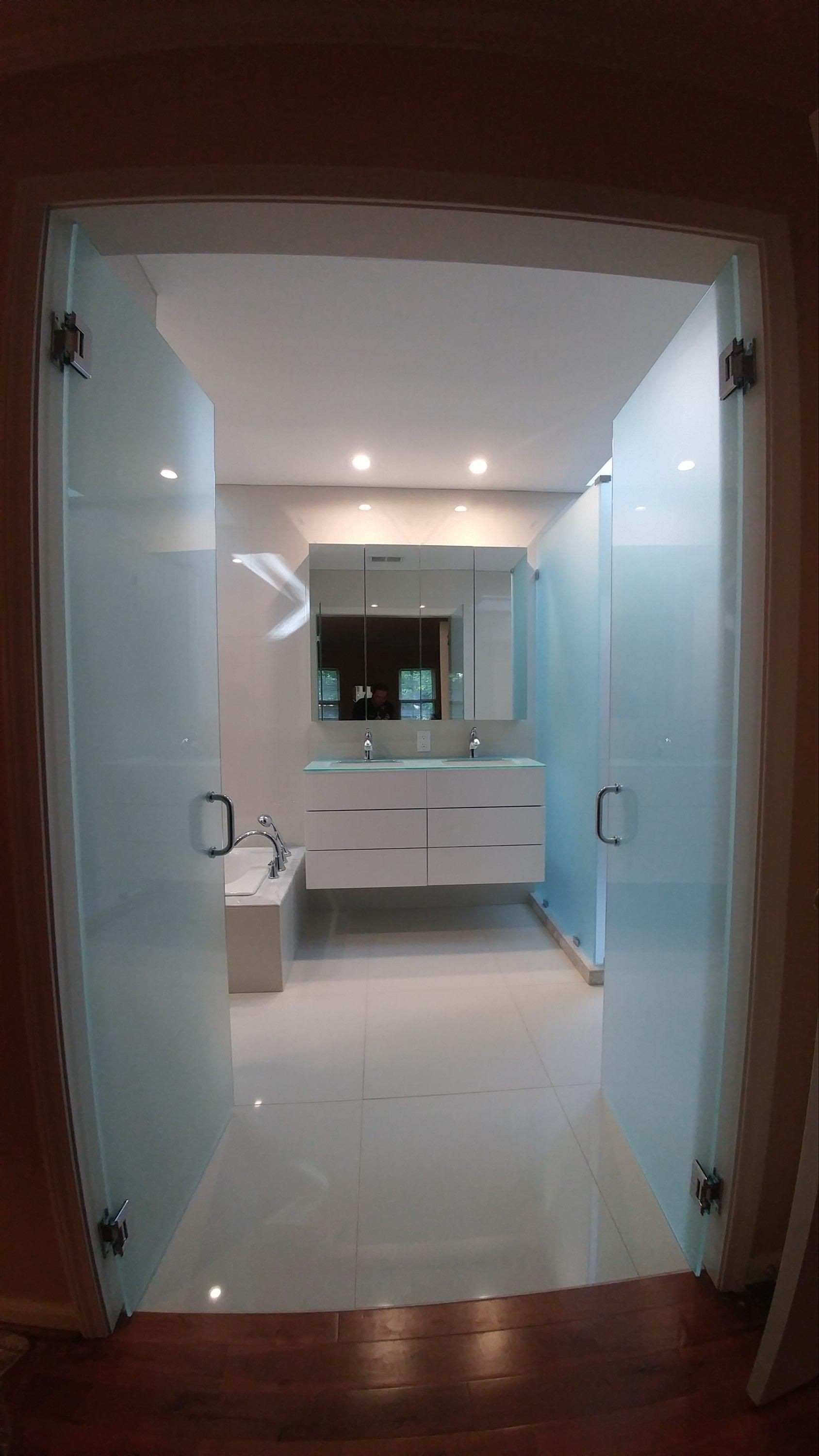 Open bathroom doorway with frosted glass doors, white vanity, mirror, and bright recessed lights.