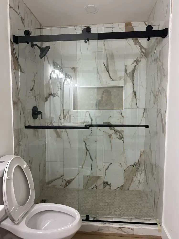 Accessible shower stall with glass door, black hardware, and marble-pattern tile beside a toilet