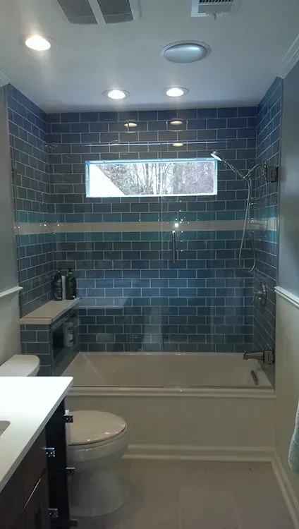 Small bathroom with blue tiled shower, narrow window, toilet, and sink countertop.