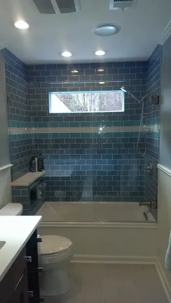 Small bathroom with blue tiled shower, narrow window, toilet, and sink countertop.