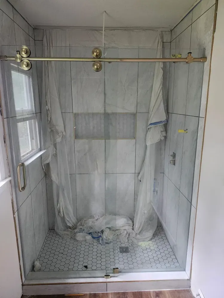 Empty tiled shower stall with marble-look walls, glass door, and brass shower rod over a floor drain.