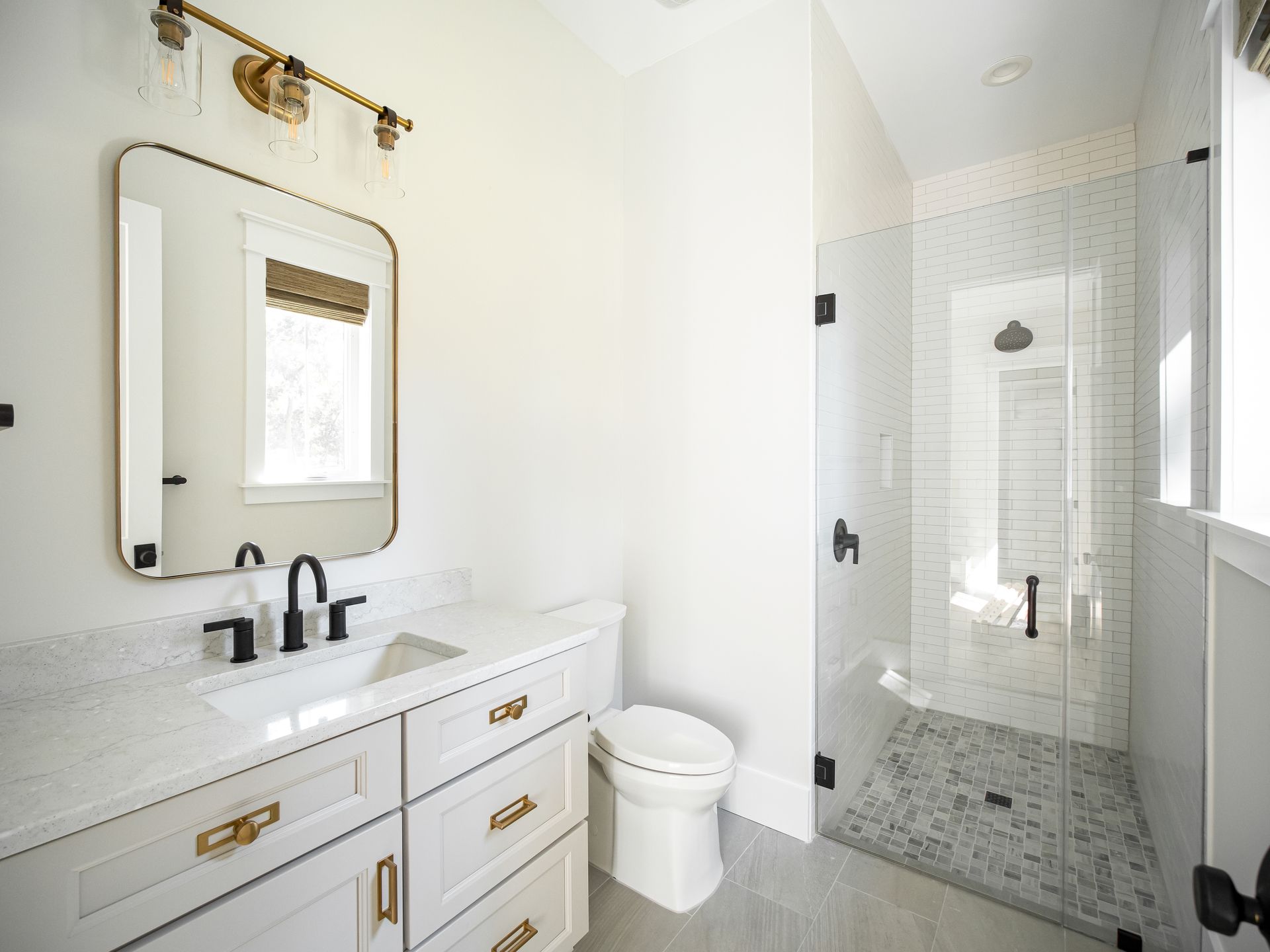 Bright white bathroom with vanity, mirror, toilet, and glass-enclosed shower with gold fixtures