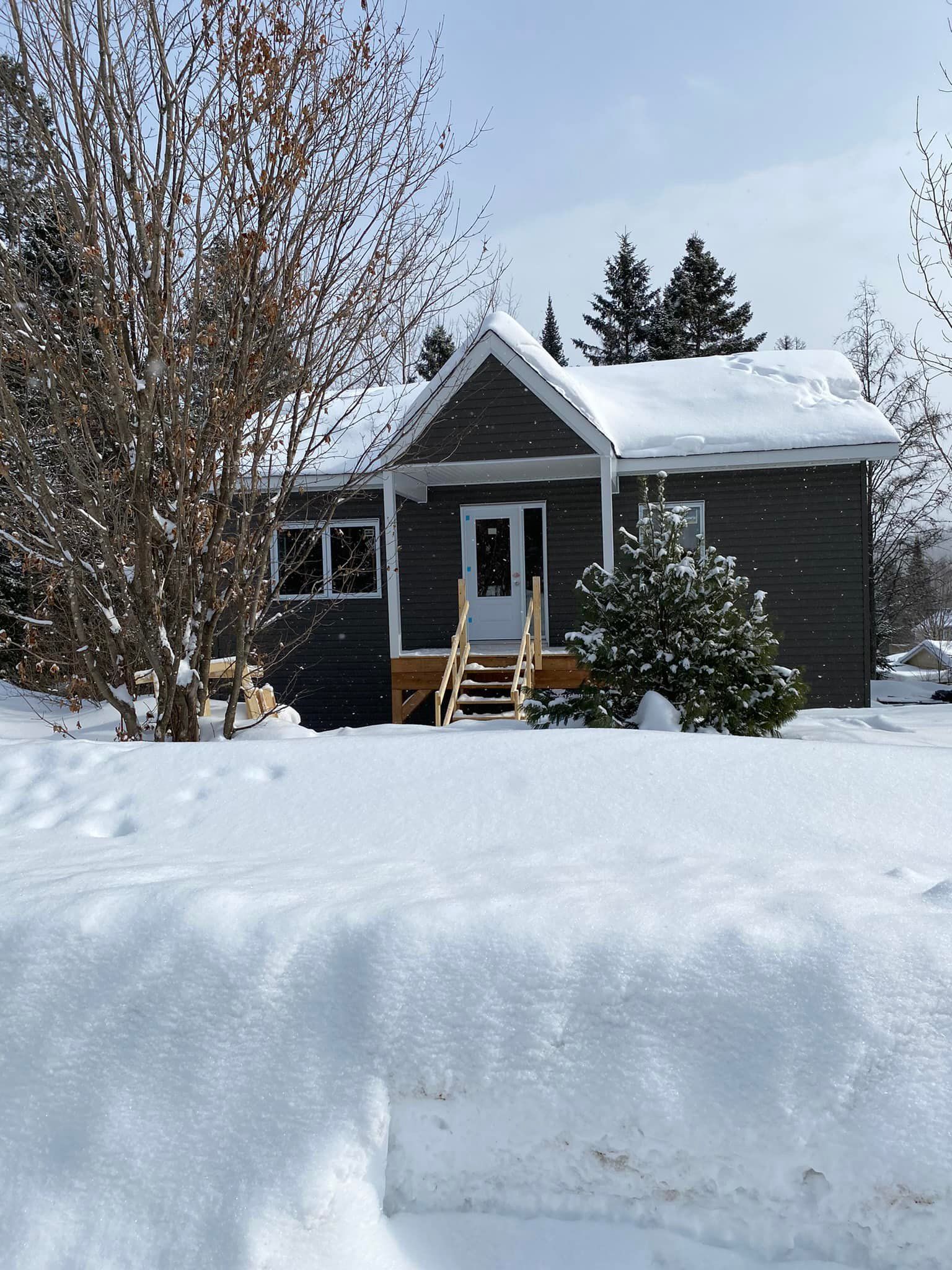 Petite maison gris foncé recouverte de neige, avec porche et escalier en bois, située dans un paysage enneigé.