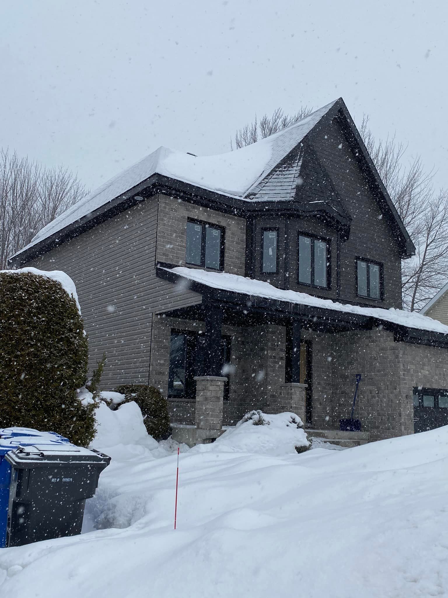Maison à deux étages avec façade en briques grises, toit enneigé et flocons de neige qui tombent.