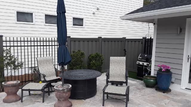 Patio with chairs, fire pit, and umbrella, next to a house with black fence and grey siding.