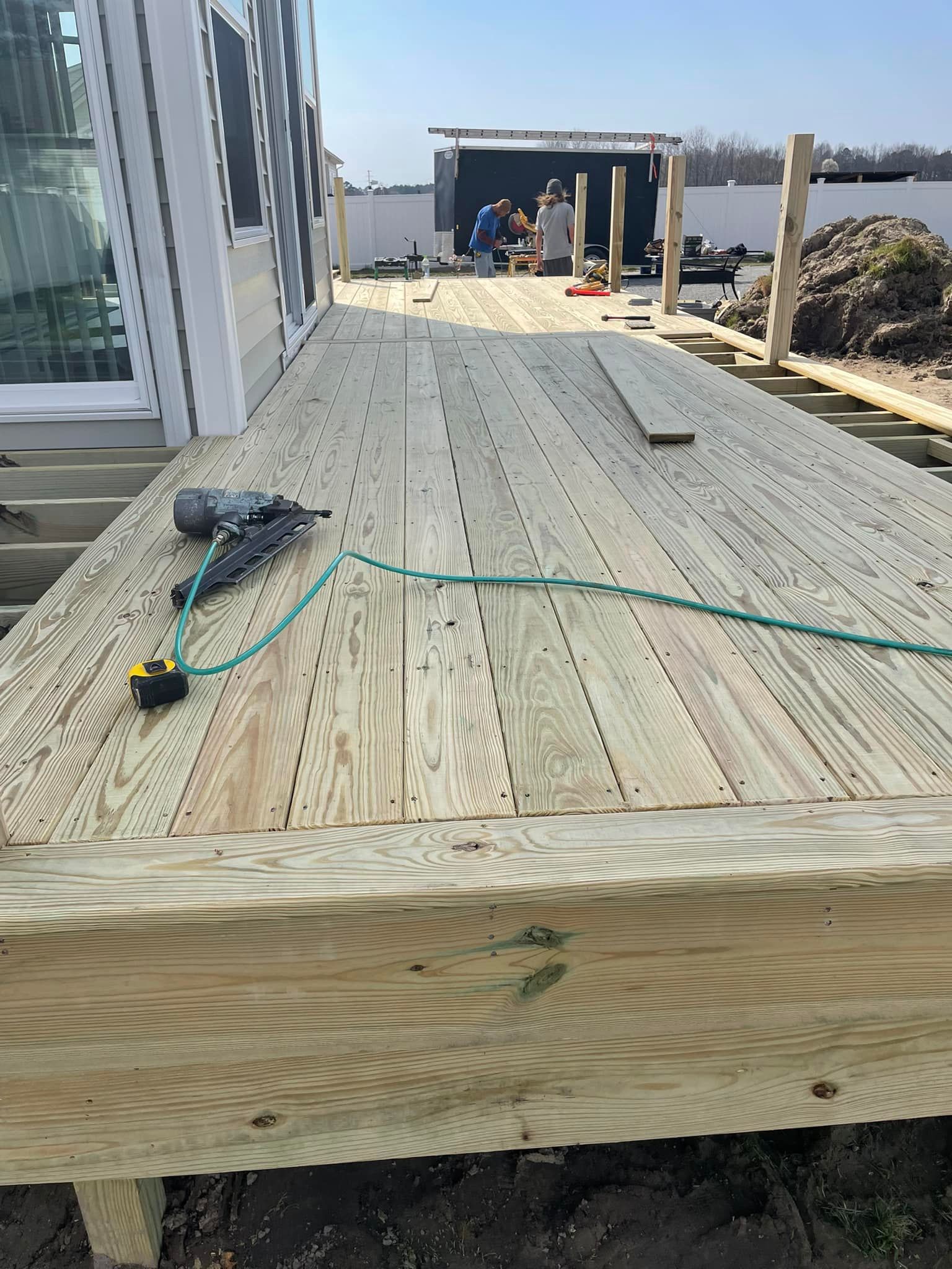 A wooden deck under construction with tools, workers in the background on a sunny day.
