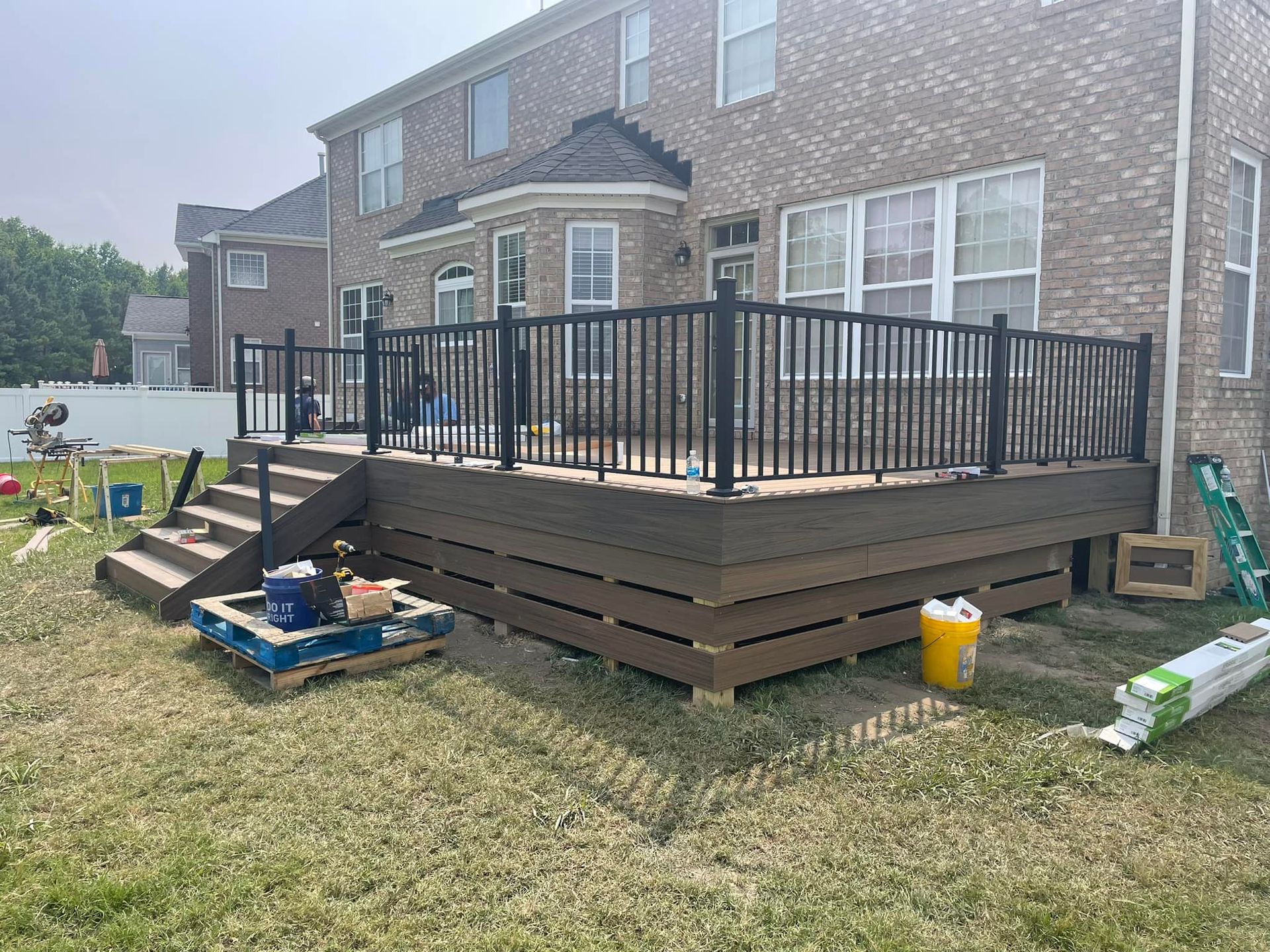 A partially constructed brown composite deck with black railing, steps, and tools scattered around a grassy yard.