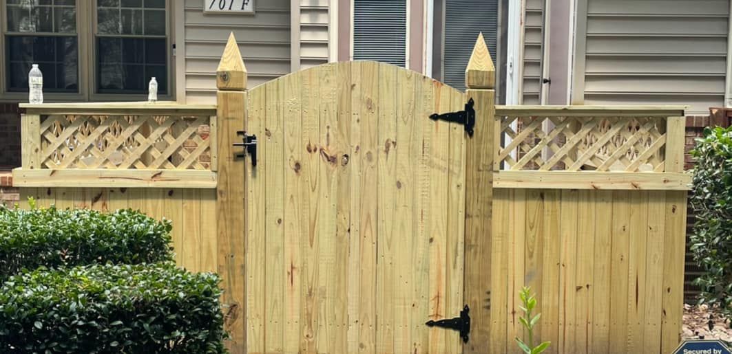 A wooden gate with pointed tops is set into a wooden fence; a house is in the background.