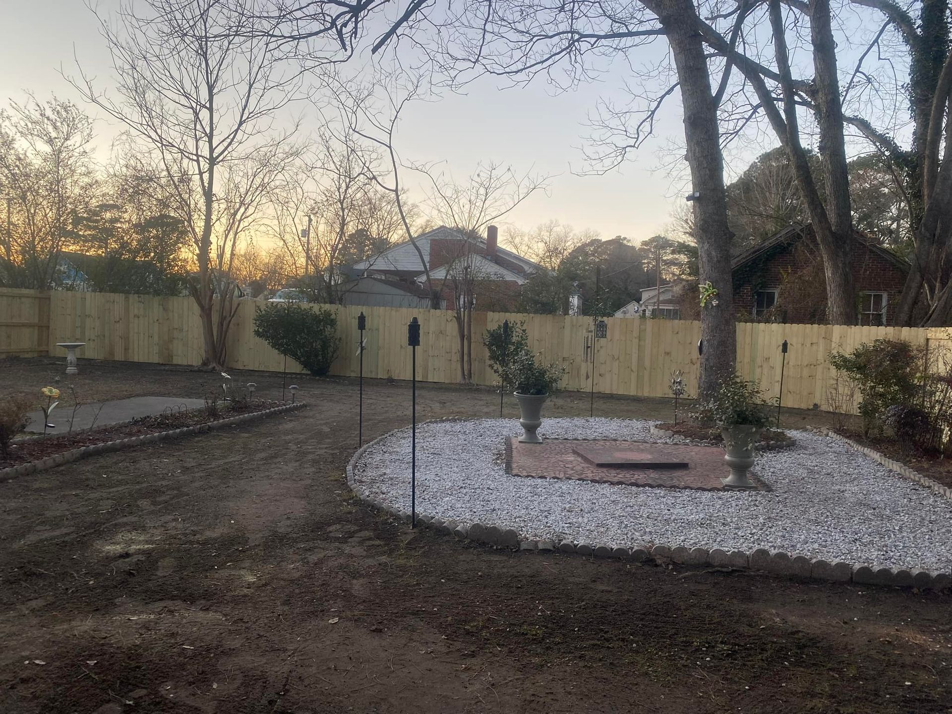 Backyard with a white rock patio, a wooden fence, and trees in the background at dusk.