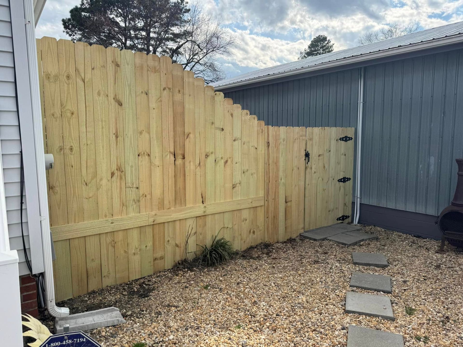 Wooden fence with a gate, gravel, and stepping stones next to a blue building.
