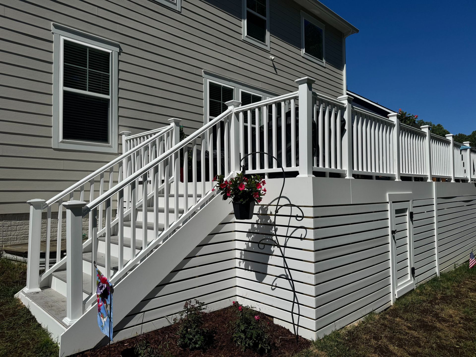 White deck stairs with railing on a gray house, with hanging flowers and a fenced yard.