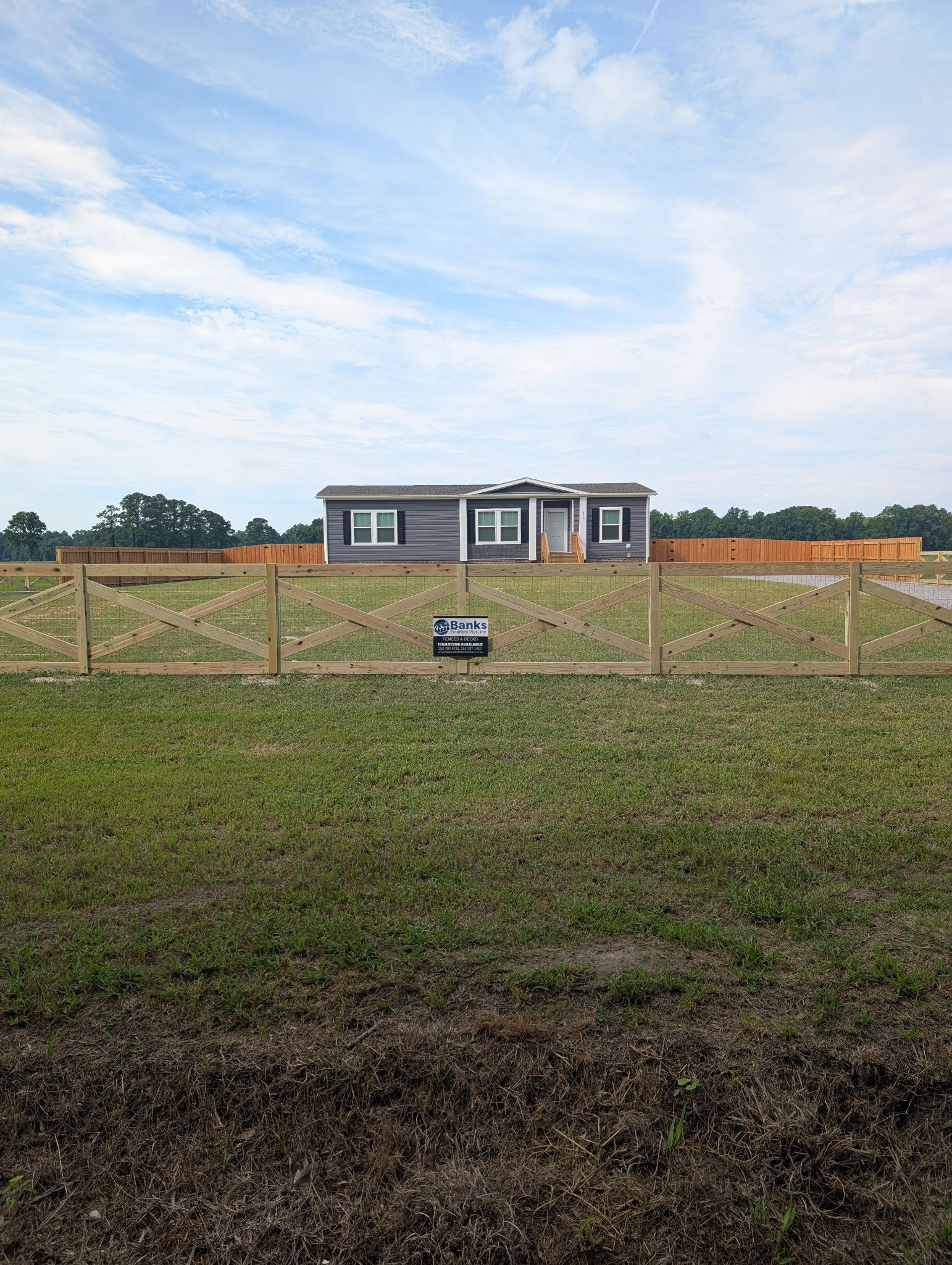 Wide grassy field with a small house in the distance under a cloudy sky, viewed through a wire fence.