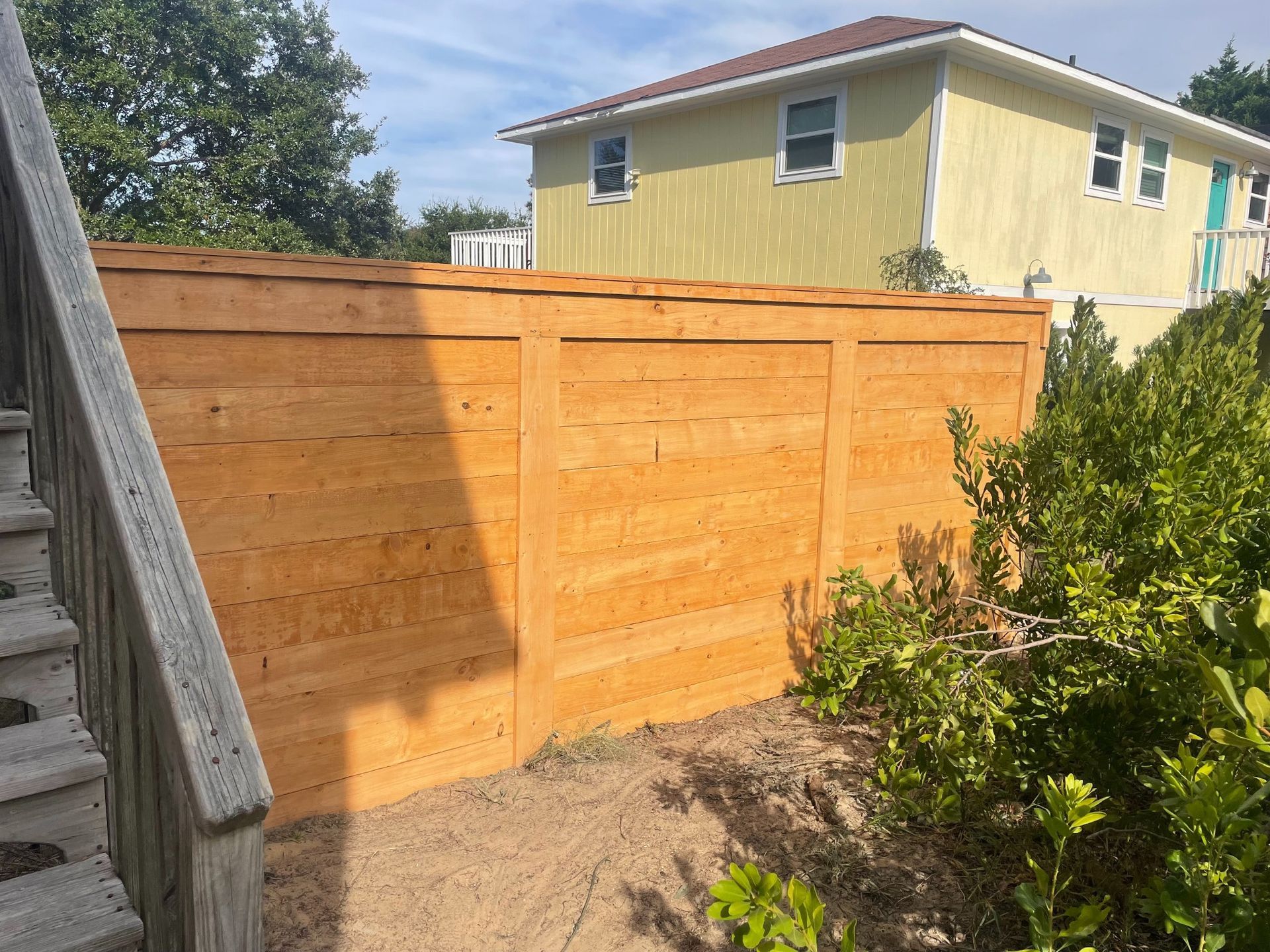 Wooden fence in front of a yellow house, blocking a view, on a sunny day.