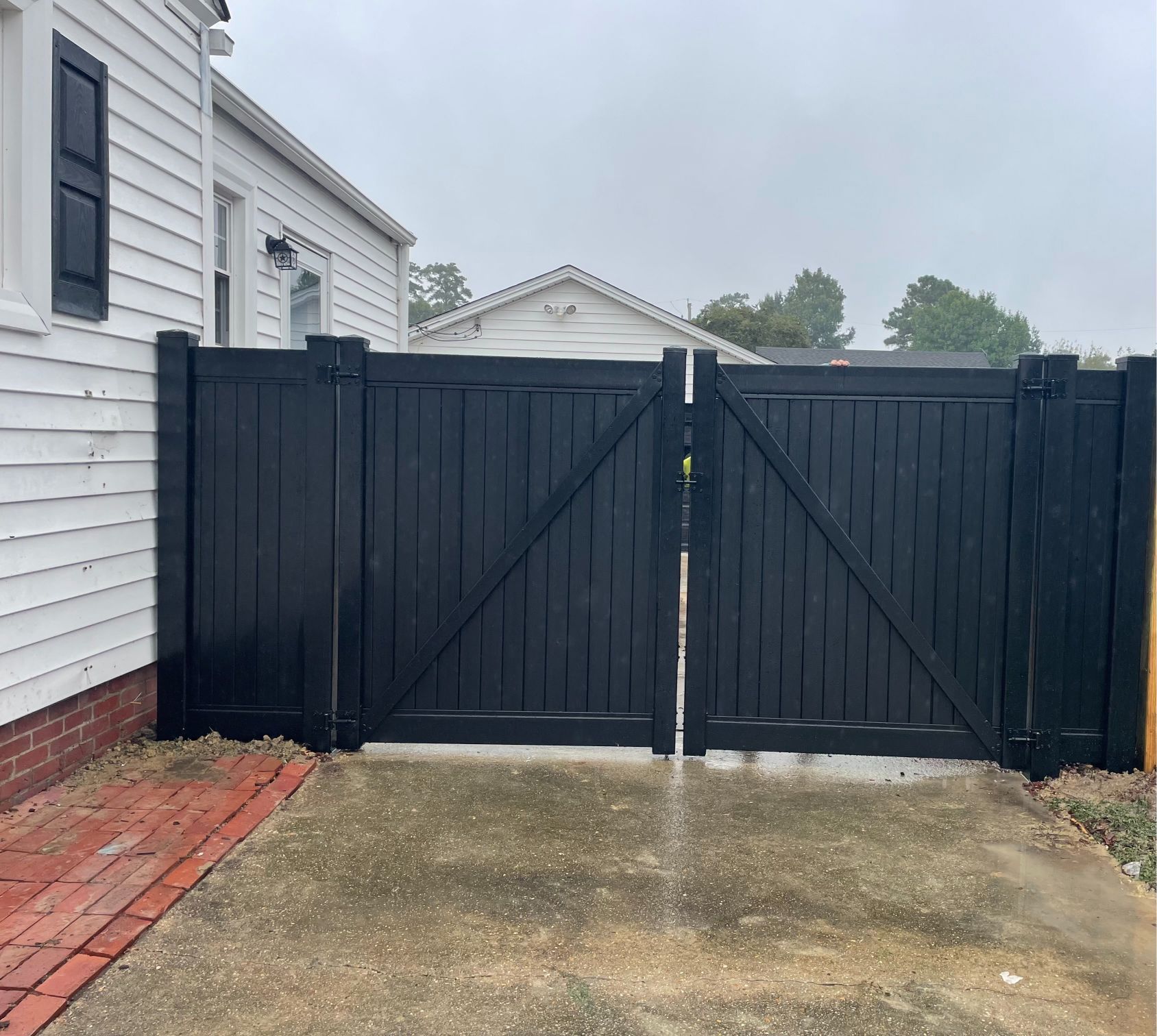 Black wooden gate in a yard next to a white house on a cloudy day.
