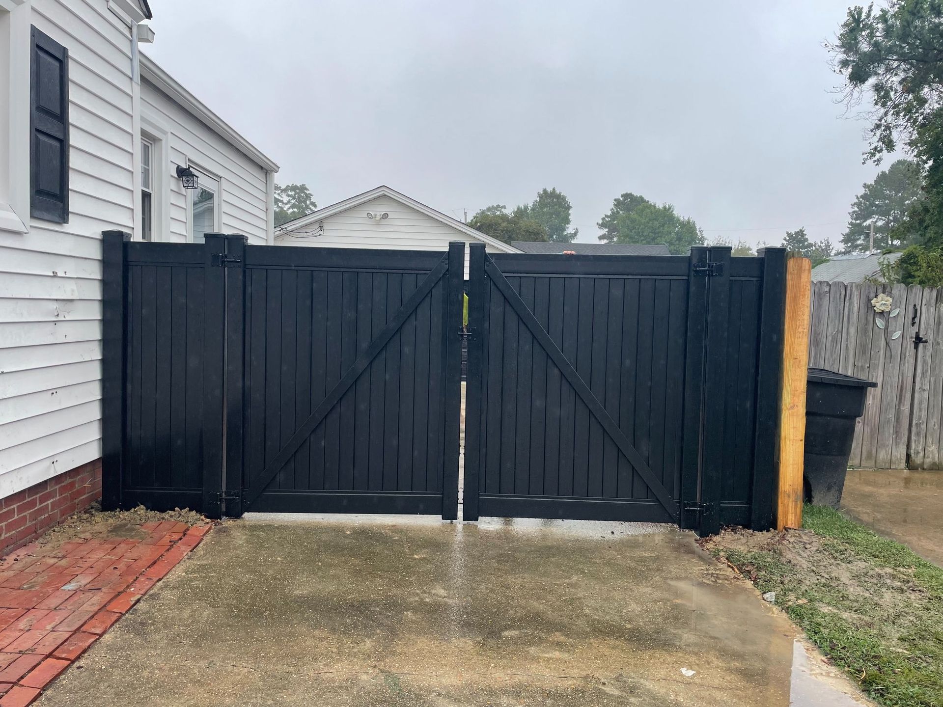 Black wooden double gate in front of a white house, set on concrete, with brick pathway.
