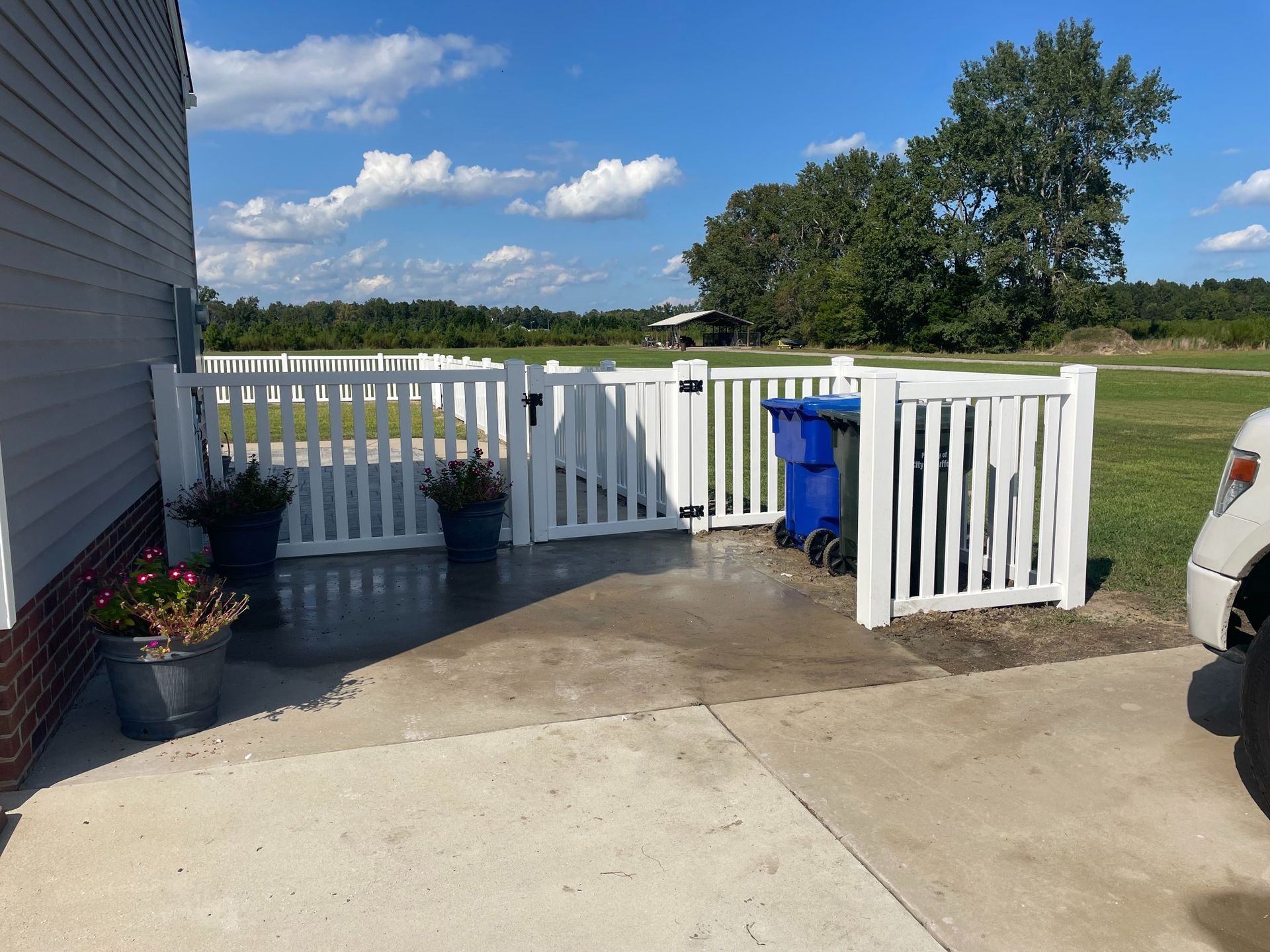 White picket fence encloses trash bins on concrete pad next to a house. Sunny day.