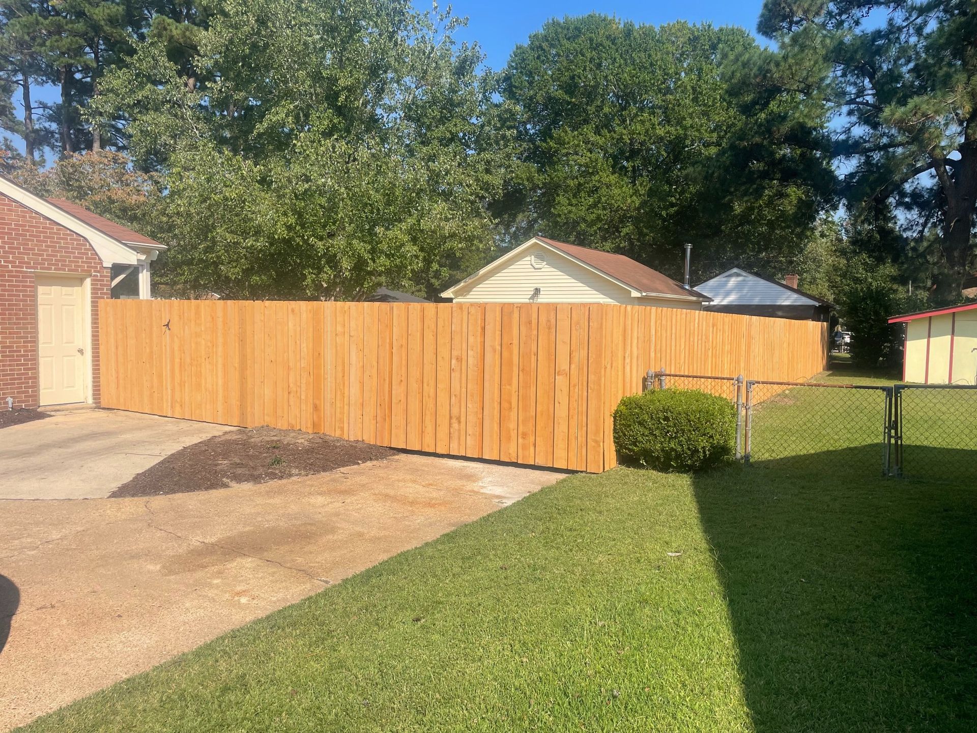 A freshly built, light-brown wooden fence encloses a yard. Green grass and trees surround it under a blue sky.