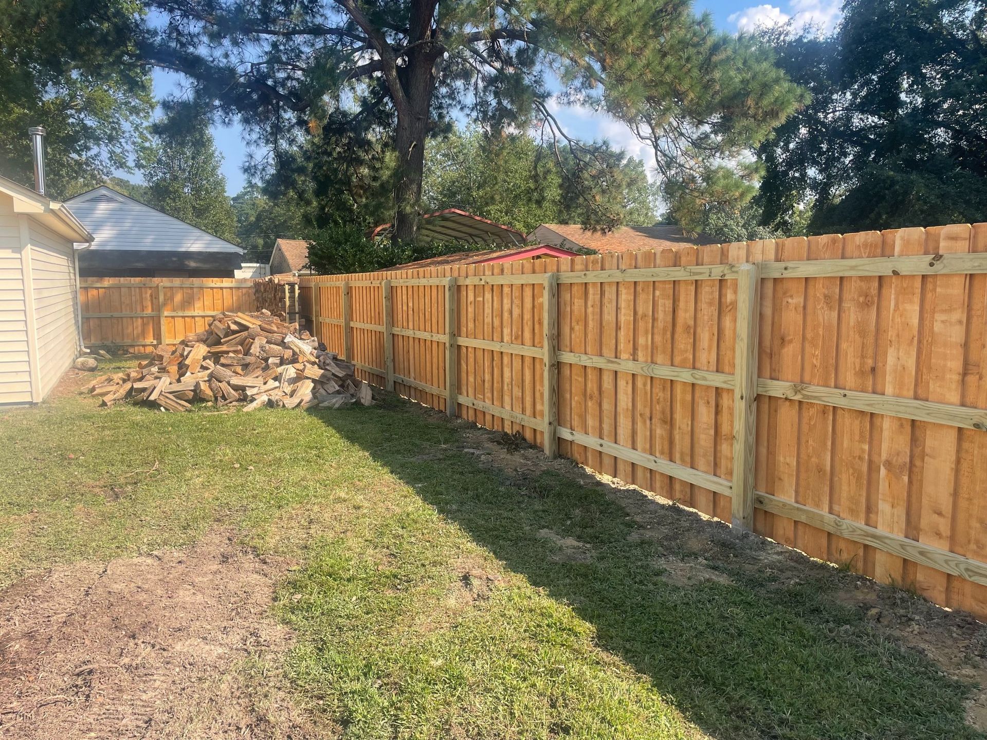 Wooden fence along a grassy backyard with a pile of wood and a house in the background.