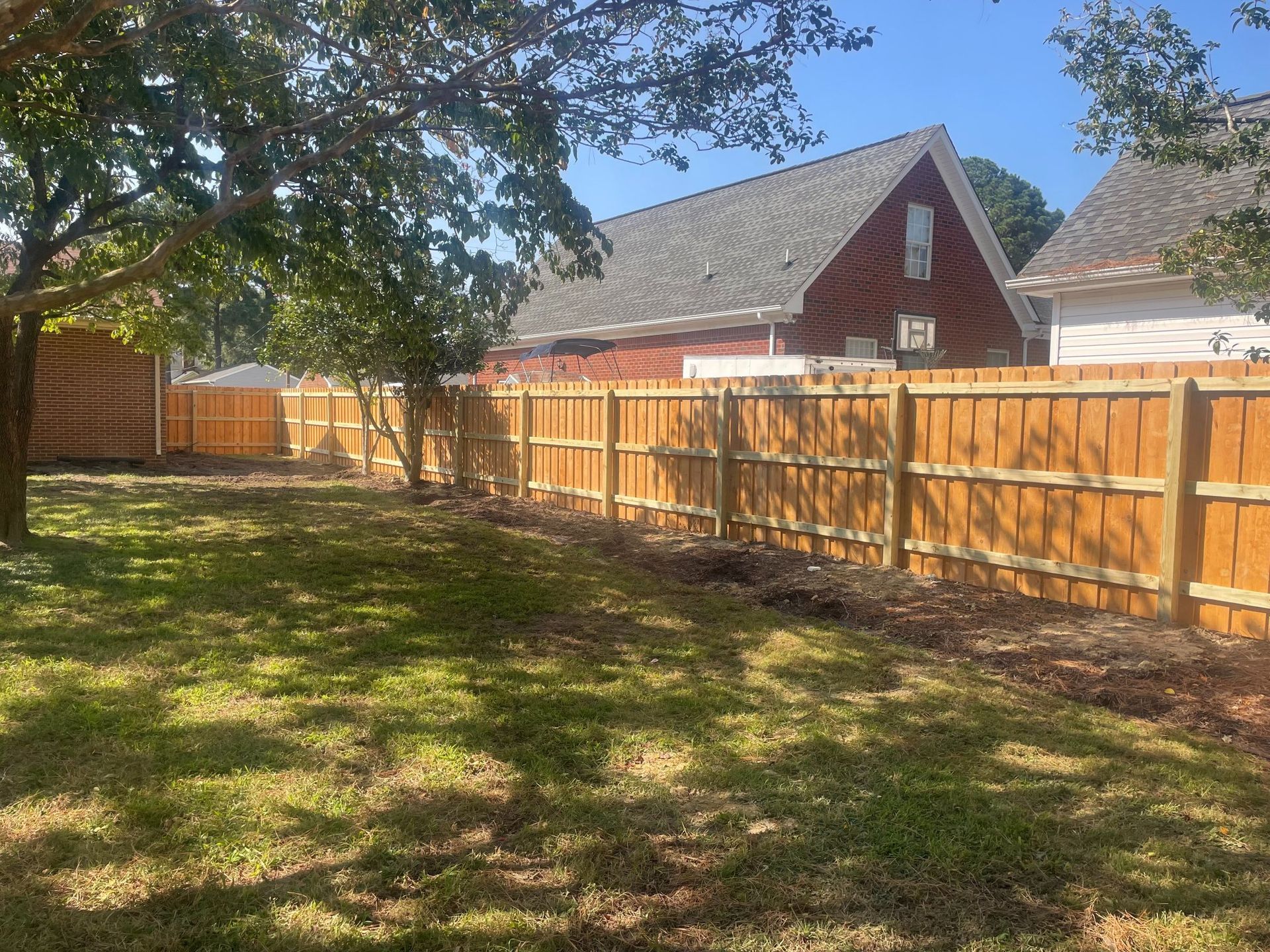 Backyard with a new wooden fence surrounding the perimeter, brown with a house in the background and trees casting shadows.