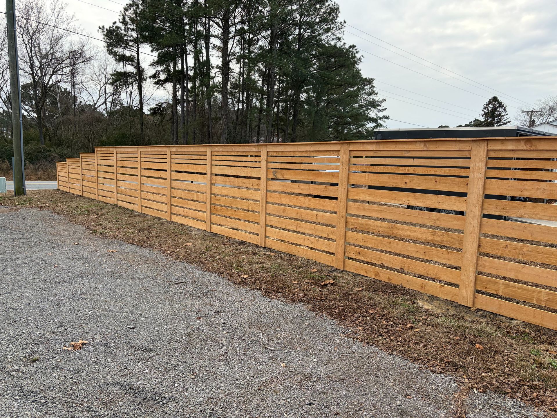 Long wooden privacy fence beside a gravel driveway, with trees and a cloudy sky in the background