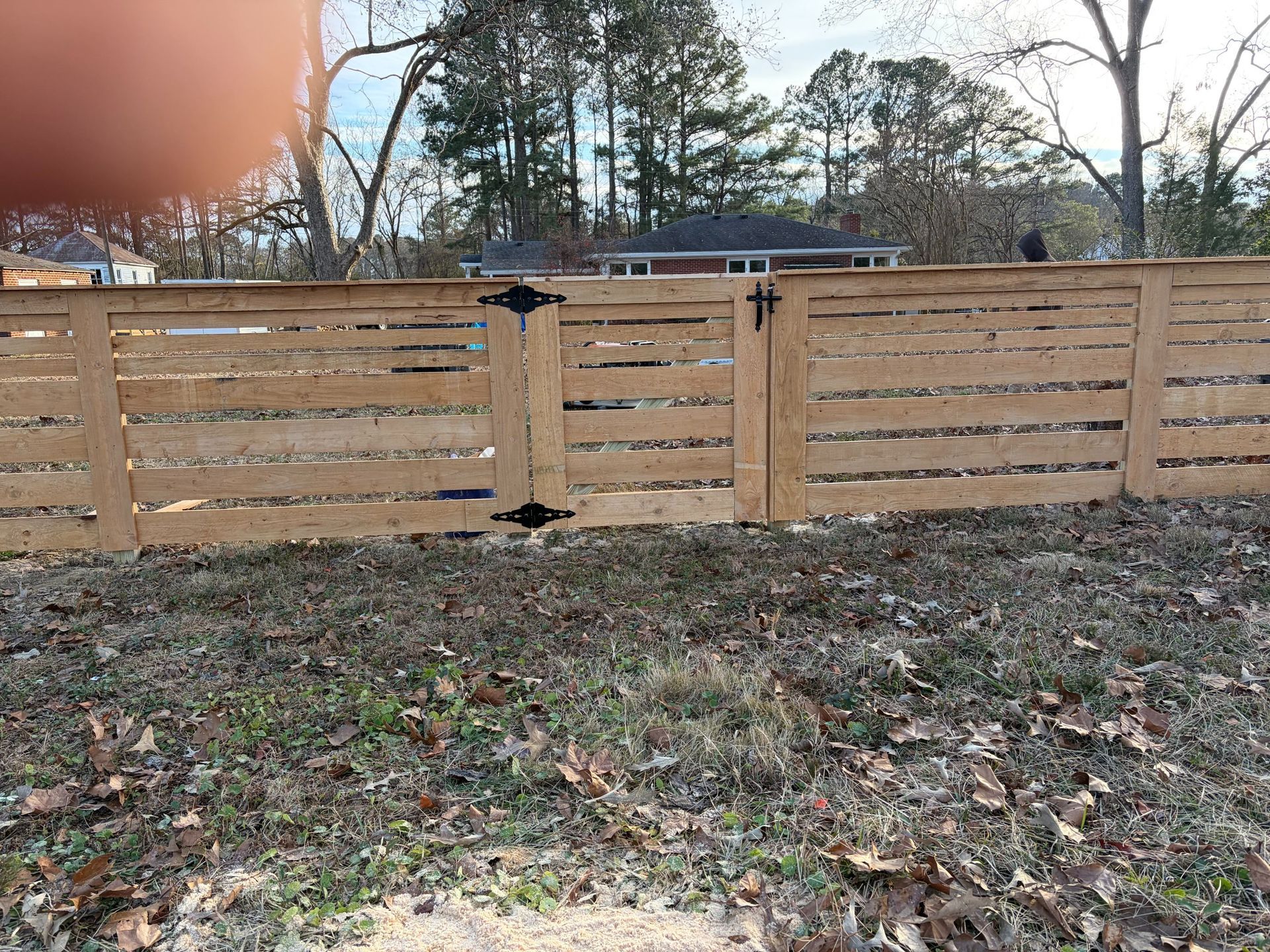 Wooden fence with gates in a leaf-covered yard, with trees and a house in the background.