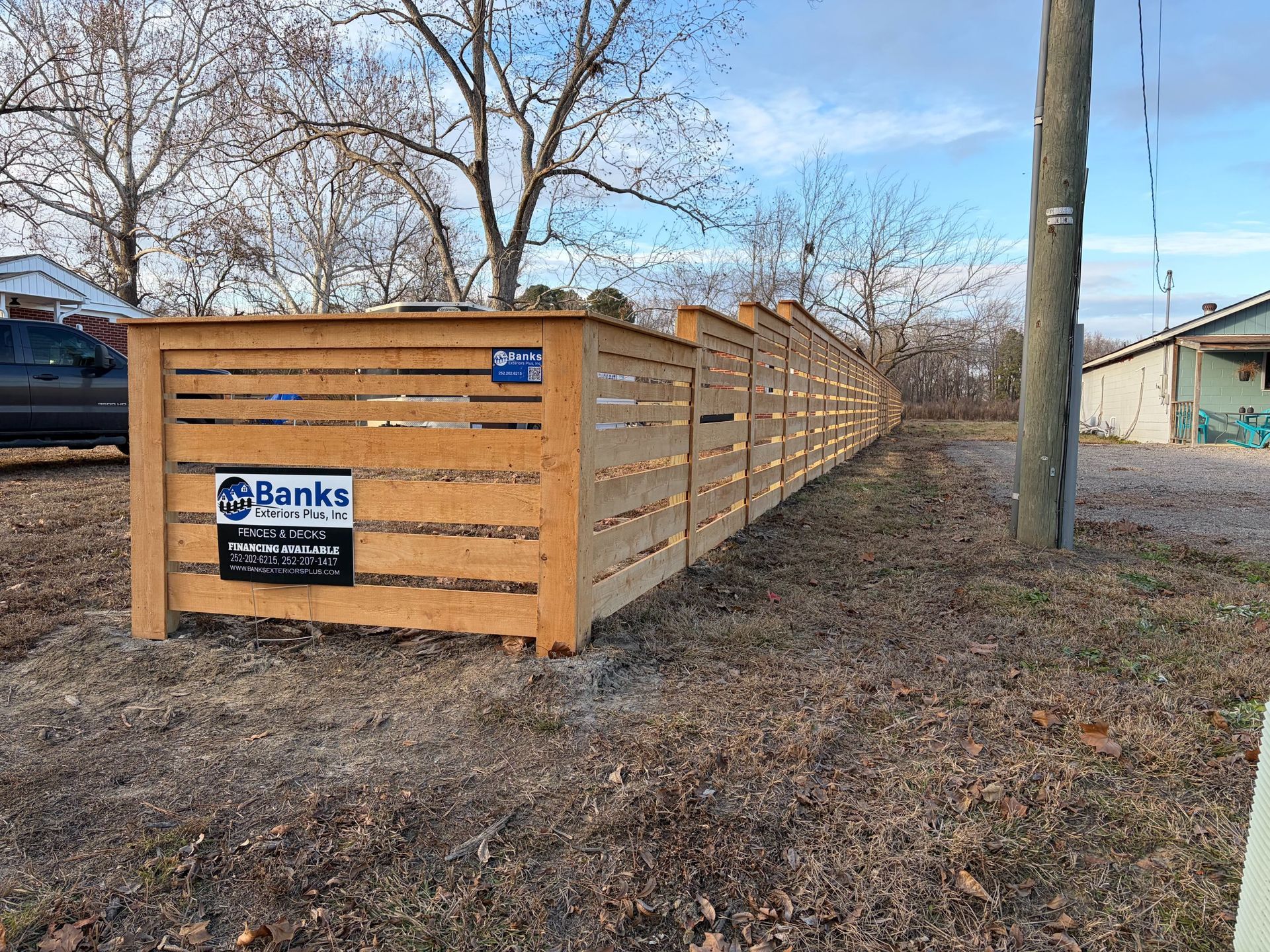 Long wooden fence panels stacked outdoors beside bare trees and a utility pole