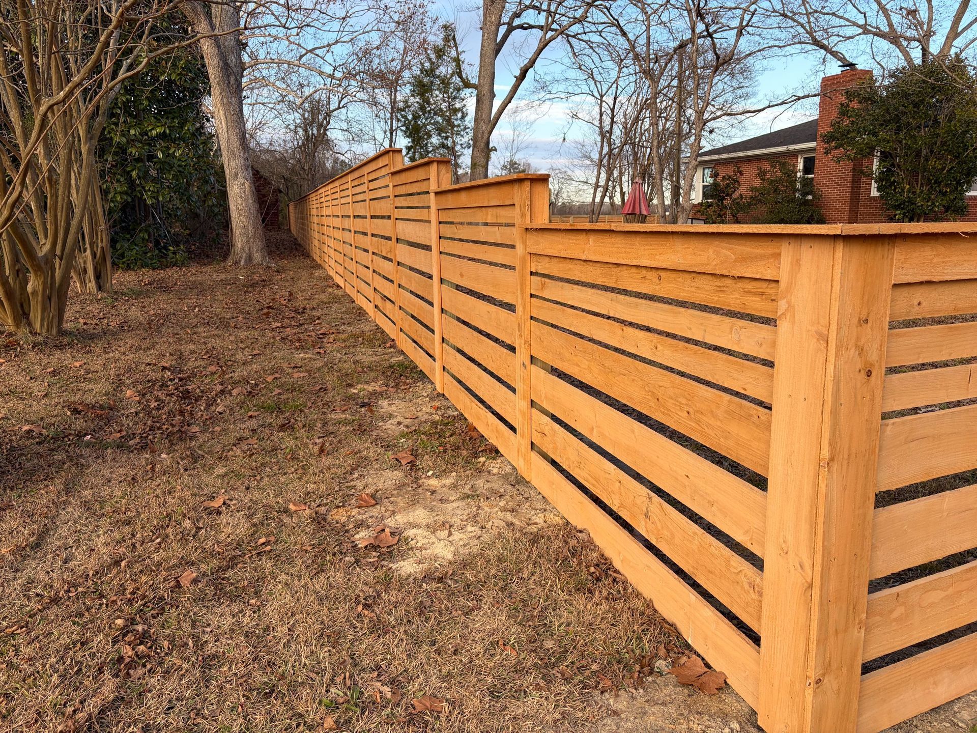 Wooden horizontal-slat fence along a leaf-covered path beside trees in a yard