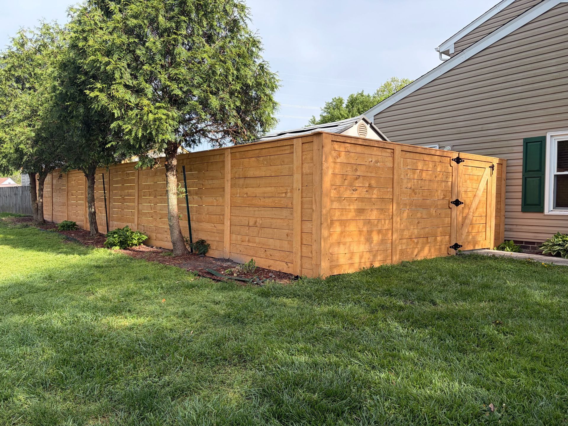 Wooden privacy fence beside a house in a grassy yard with a large tree.