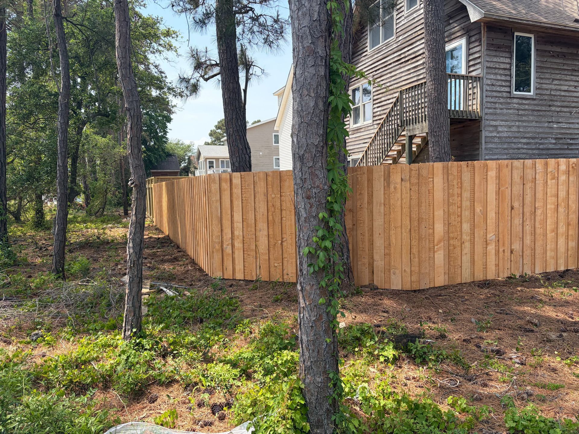 Wooden privacy fence around a house in a wooded yard with tall pine trees and stairs to a deck.