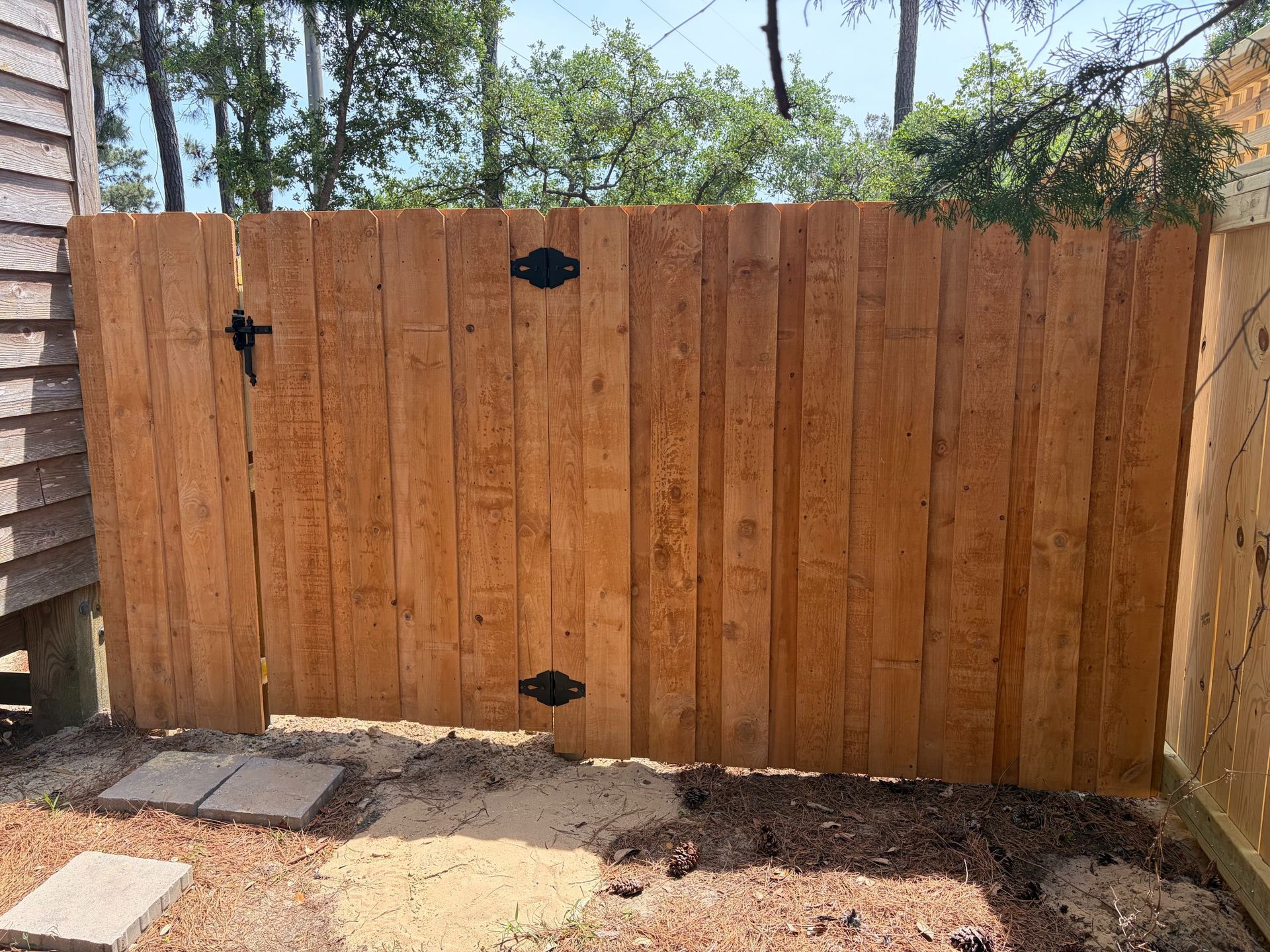Wooden backyard gate with black hinges between fences, with trees and patio stones nearby