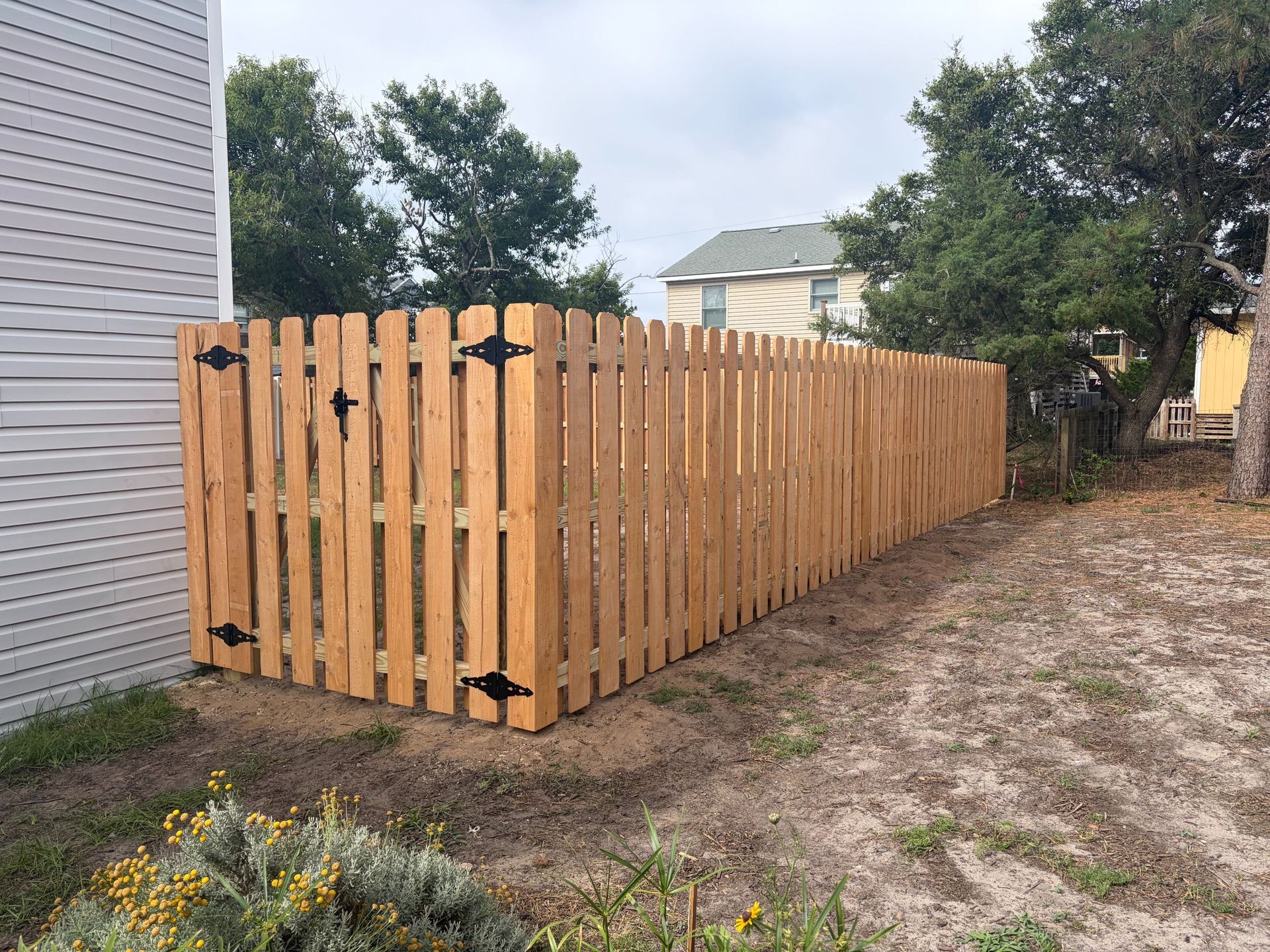 Wooden fence along a dirt path beside a house, with trees and a yard in the background.
