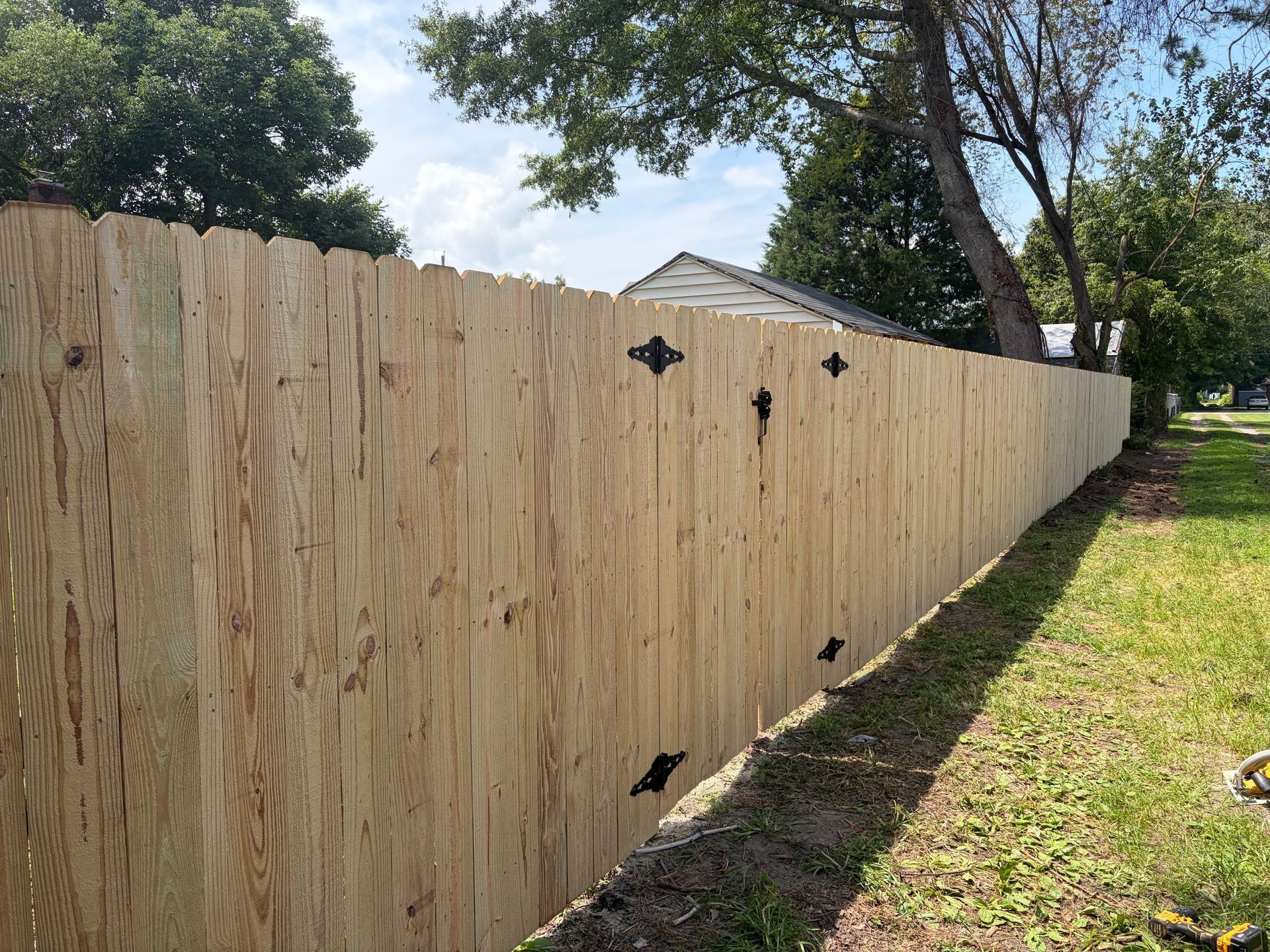 Wooden privacy fence along a grassy yard, with black hinges and trees overhead.