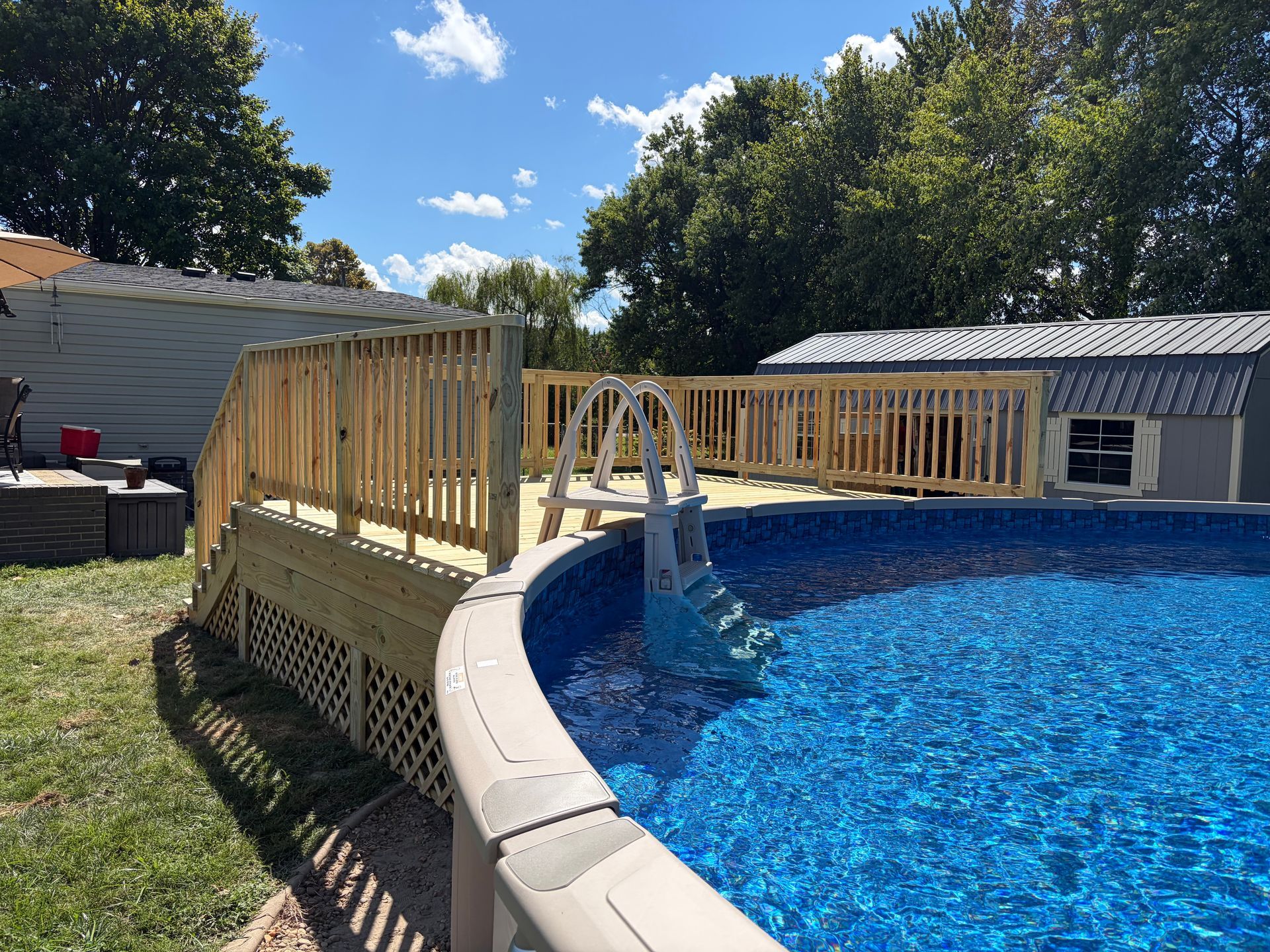 Backyard above-ground pool with a wooden deck railing and clear blue water under a sunny sky