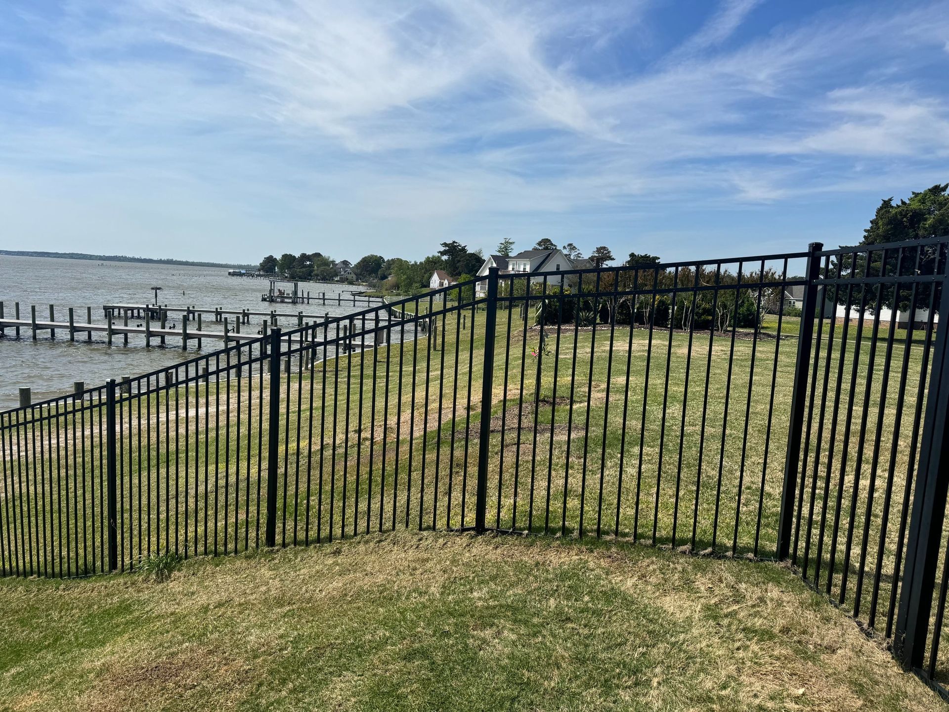 Black metal fence along a grassy waterfront path with a lake and blue sky in the background