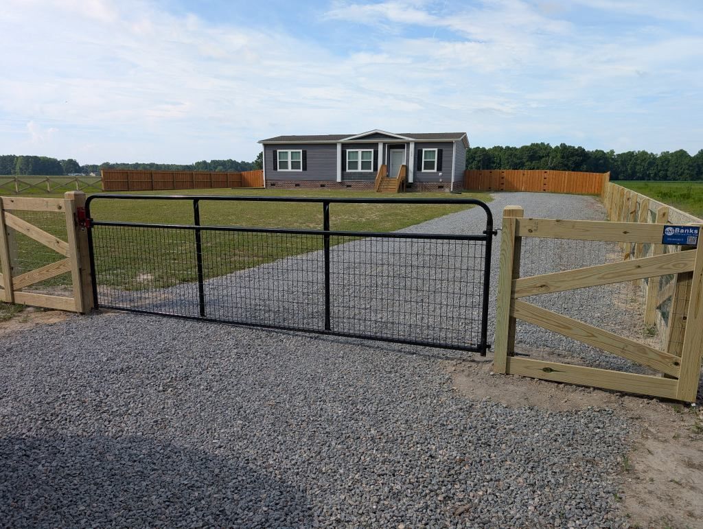 Gravel driveway with open wooden gate leading to a small house in a rural field.
