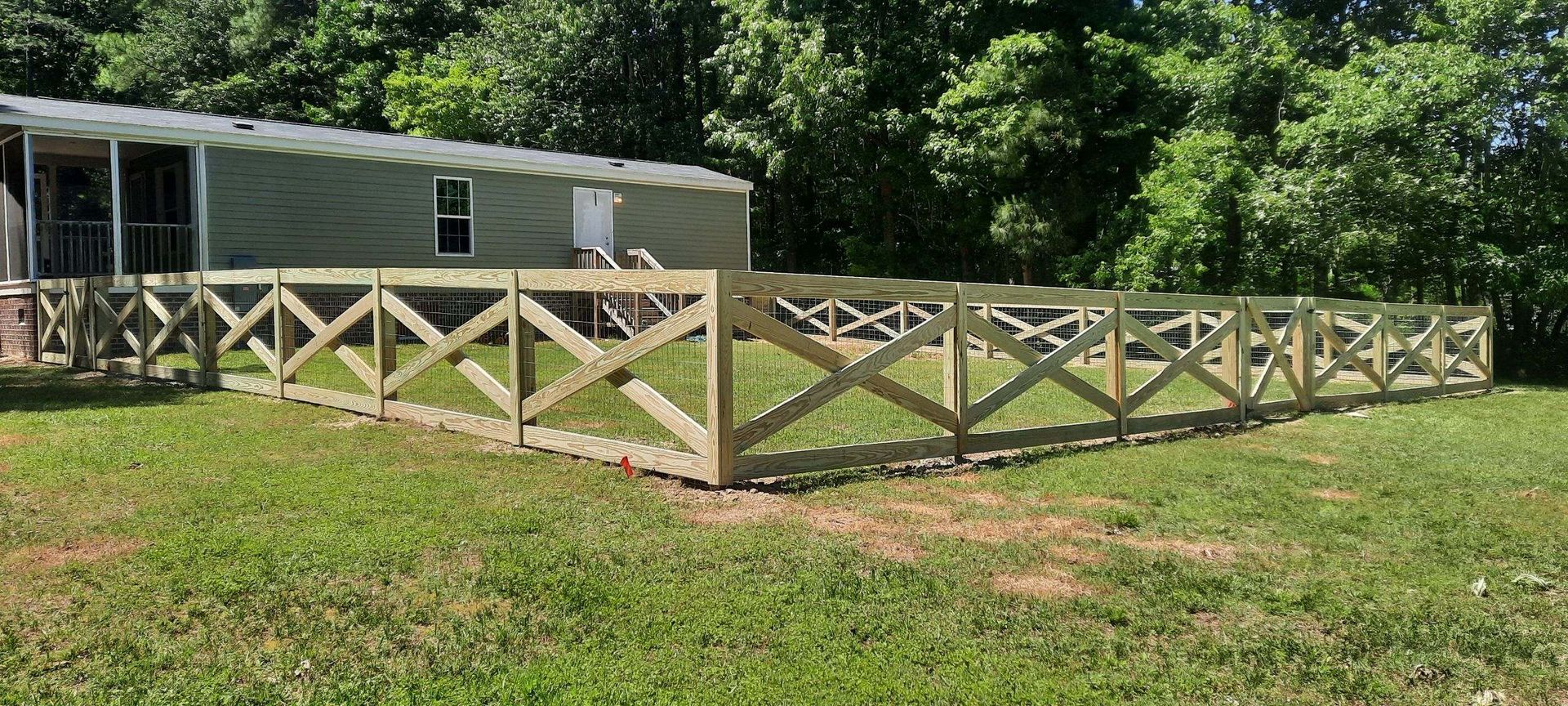 Beige fenced yard beside a small house on a sunny grassy lot with trees in the background
