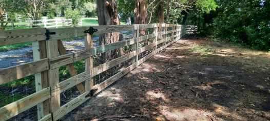Wooden fence along a shaded dirt path beside a green field and trees