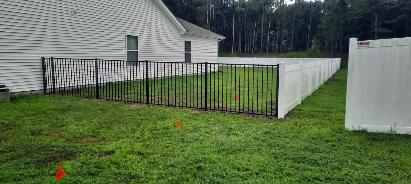 Black metal fence encloses a green lawn beside a white house and privacy fence.