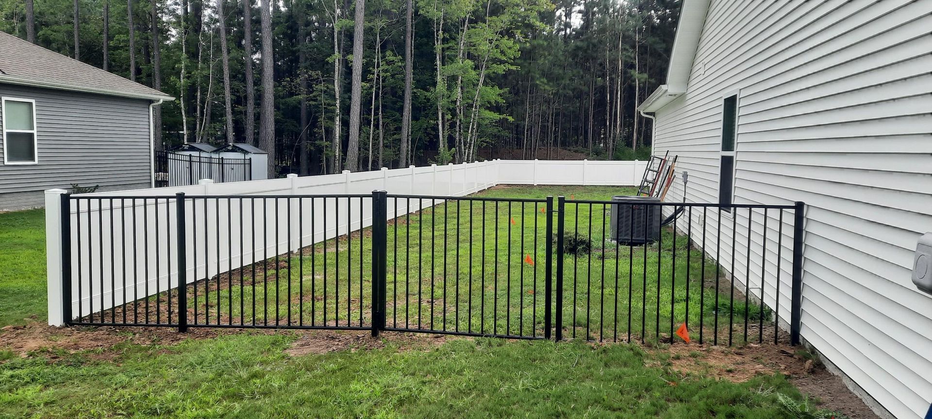 Black metal fence section between houses, enclosing a grassy backyard with a white fence gate and lawn chairs.