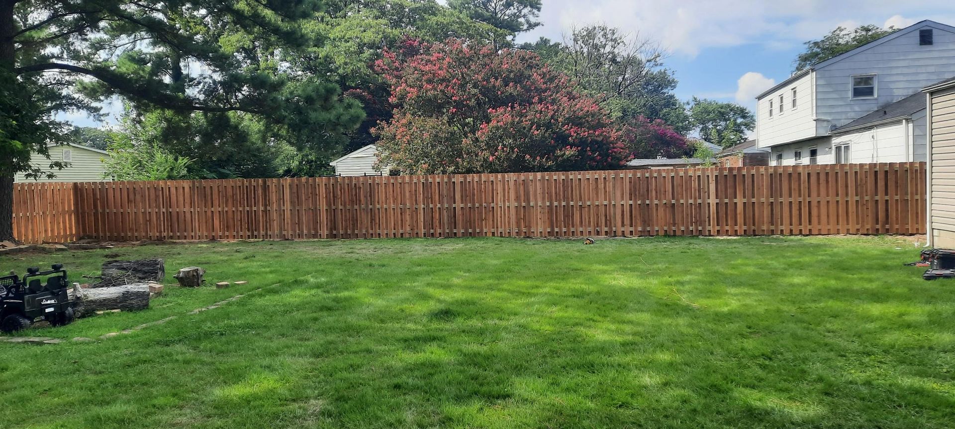 Backyard with green lawn, wooden privacy fence, trees, and neighboring houses under a partly cloudy sky