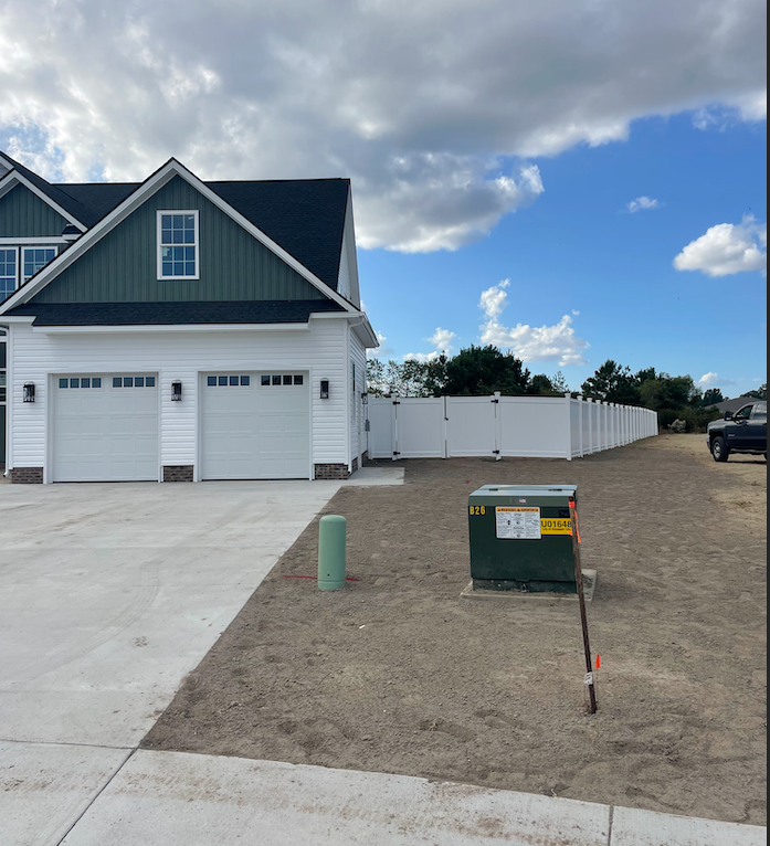Two-car garage attached to a white house, with a white fence and a utility box in a yard with dirt.