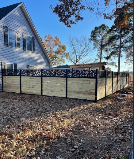 A wooden fence with a decorative top, in front of a house, set in a grassy yard.