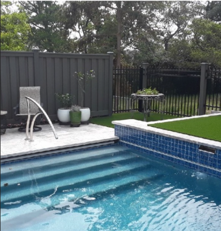 Pool with blue tile, steps, and water, adjacent to a patio with a chair, plants, and a dark fence.