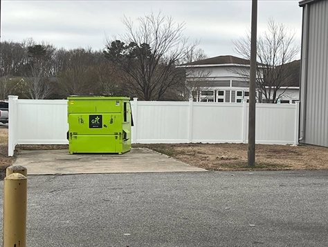 Bright green dumpster behind a white fence, beside a building on a cloudy day.
