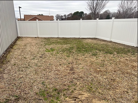 A small, fenced backyard with dead grass and a cloudy sky. White vinyl fence and building on left.