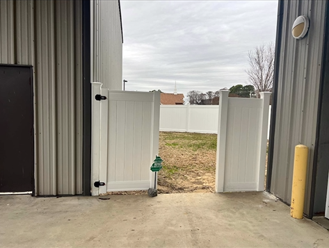 White fenced gate open between metal buildings, with a grassy yard beyond.