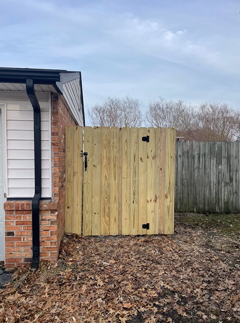 Wooden gate attached to a brick building, opening to a yard with a fence. Dry leaves cover the ground.